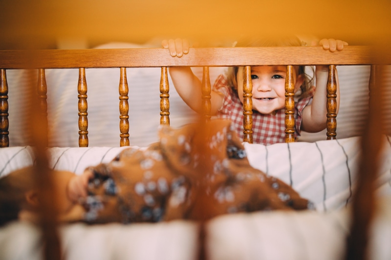 Fresh 48 Newborn Photography, a little girl peaks over a wooden crib to admire her newborn sibling sleeping, she smiles