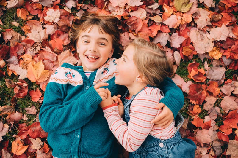 Family Photography - two young sisters laugh and hold each other close as they lay in the fall leaves outdoors
