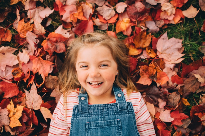 Family Photography - a little girl lays in the autumn leaves in the grass smiling