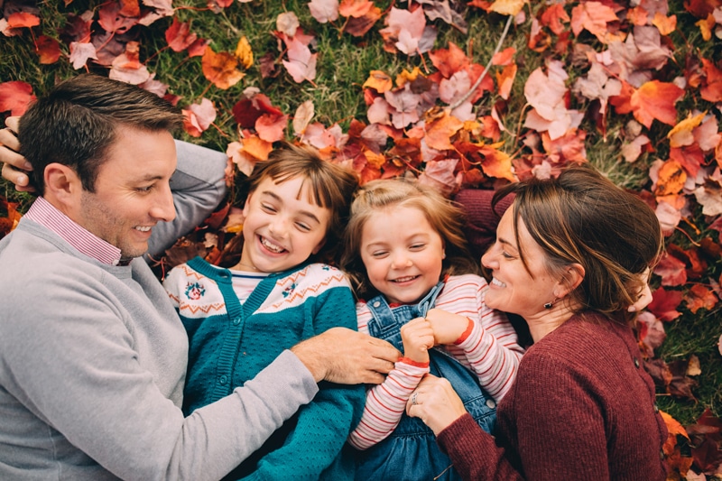 Family Photography - a young family of four laugh as they lay in the fall leaves and hold each other close