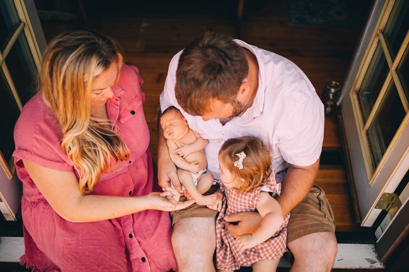 Fresh 48 Newborn Photography, a dad holds baby and older sister in his lap, mom admires them