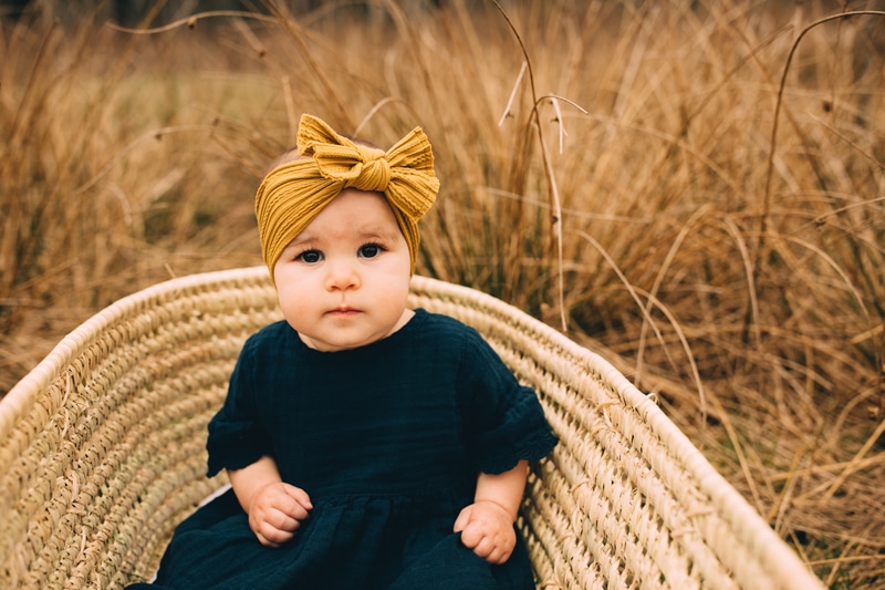 Milestone Photography, a baby girl sits in a woven basket outside in a dry brush