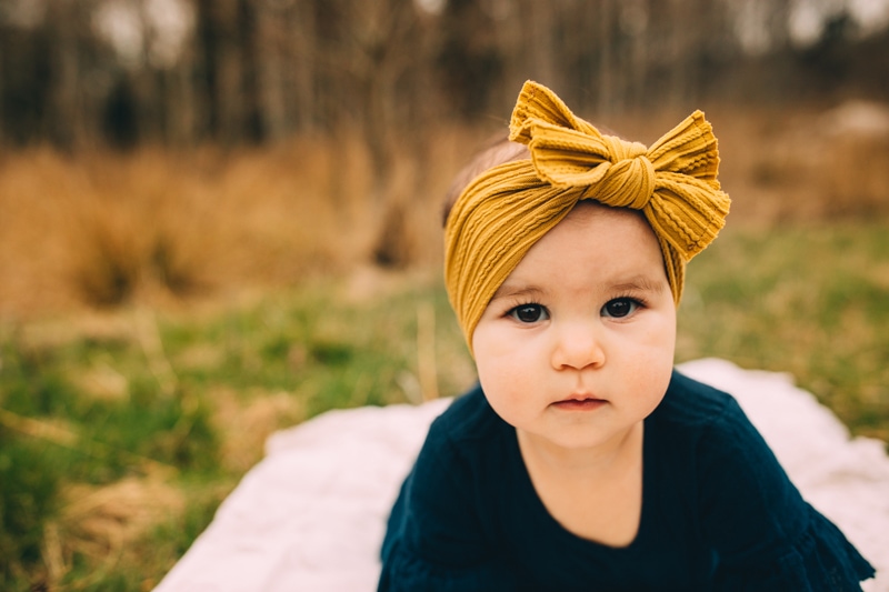 Milestone Photography,  a baby girl sits on a. picnic blanket outside in wooded area with a big bow wrapped on her head