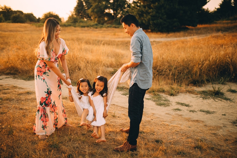 Family Photography - a mother and father hold a sheet and swing their two young daughters, all smiling, they are in a dry grassy field outdoors