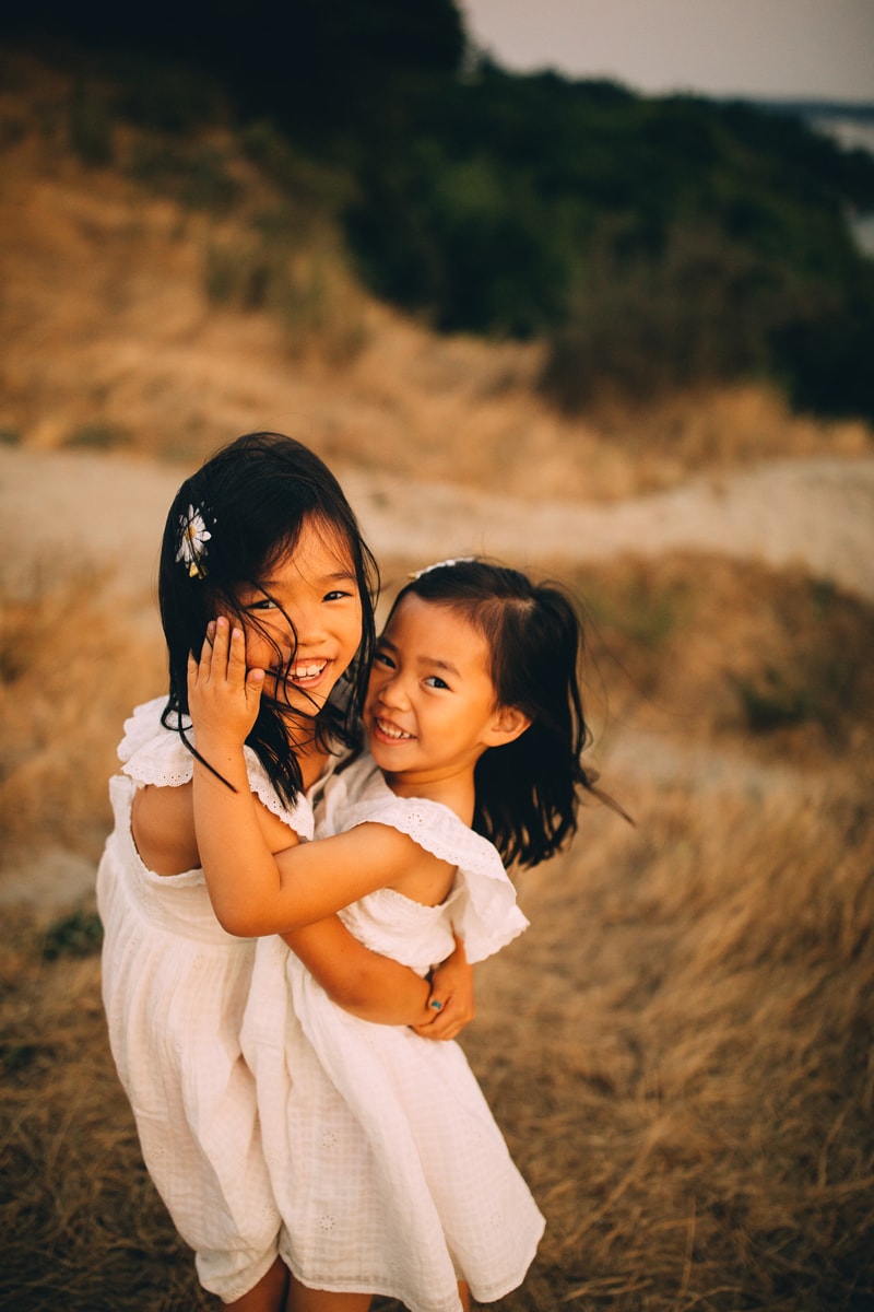 Family Photography - two young sisters embrace and smile with affection on a windy day in a dry grassy field