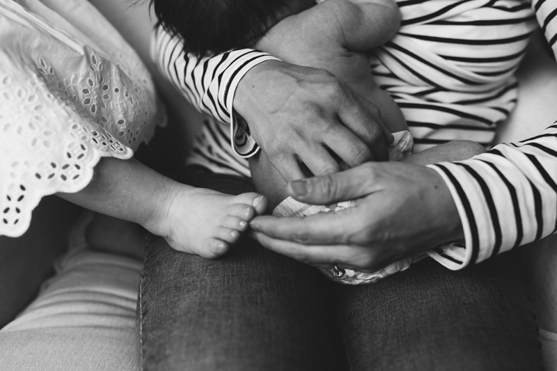 Fresh 48 Newborn Photography, a mother holds baby and older sisters toes reach out to mom's lap for comfort too
