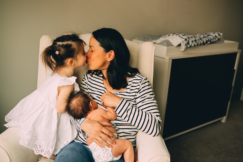 Fresh 48 Newborn Photography, a new mother sits in a chair in baby's nursery breast feeding, her other daughter reaches in for a kiss