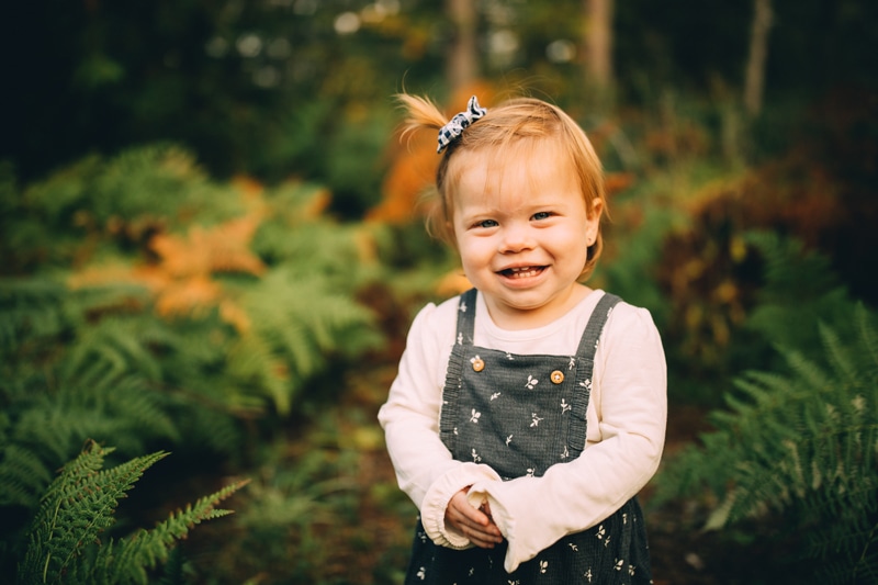 Milestone Photography, a young girl smiles as she stands among ferns in the forest