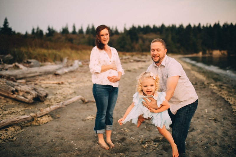 Family Photography - A dad swings his toddler girl playfully in the sand near a lake, mom looks on, pregnant and holding her belly
