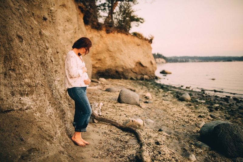 Family Photography - an expecting woman stands near a lakeshore holding her belly in anticipation