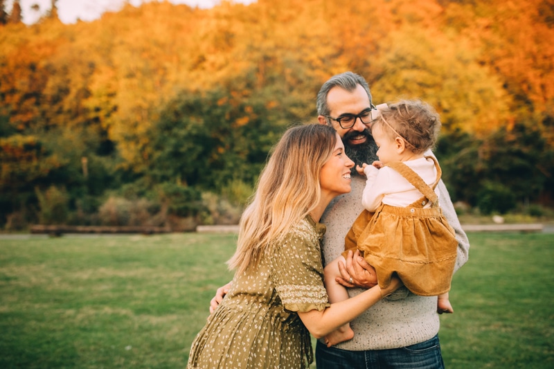 Family Photography - Dad holds daughter and mom leans in close to give her attention, They are outside in a wooded area and happy