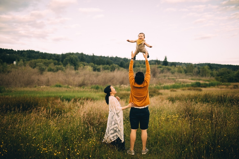 Family Photography - a father tosses baby into the air playfully in a grassy meadow, mom looks on and laughs