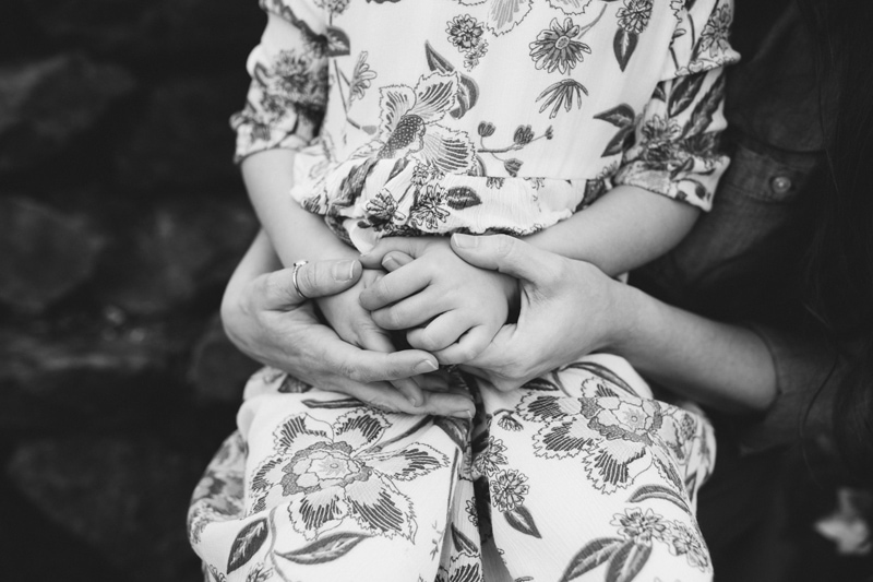 Motherhood Photography, a mother holds the hands of her daughter as she sits on her lap