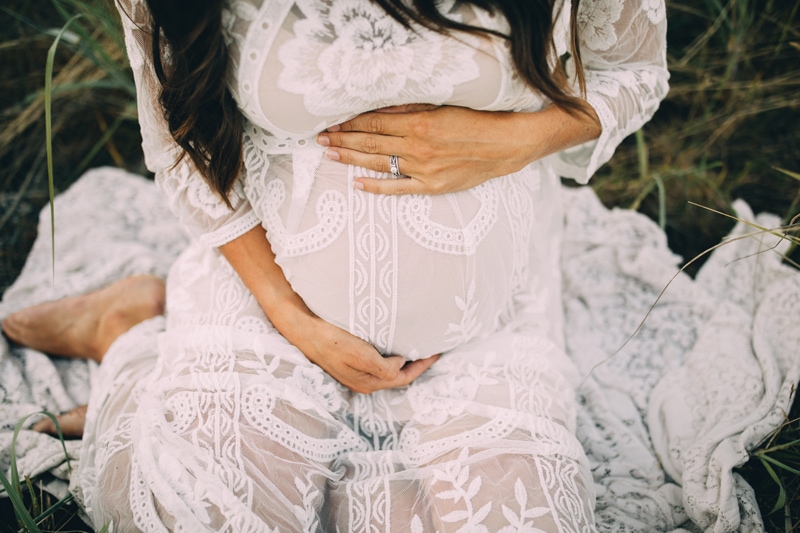 Family Photography - a woman in a white lacy dress sits in the grass and holds her belly pregnant
