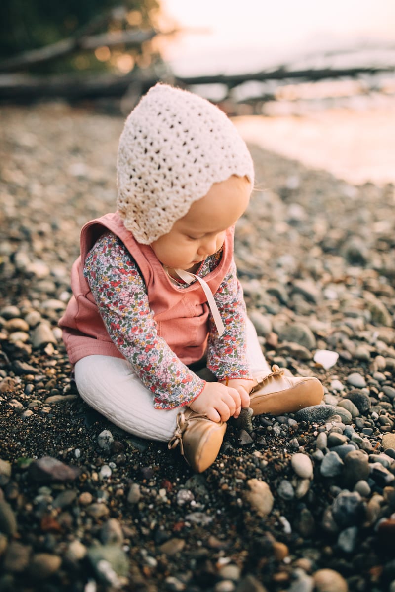 Milestone Photography, a baby girl with a knit cap sits on a lake shore curiously looking at the stones