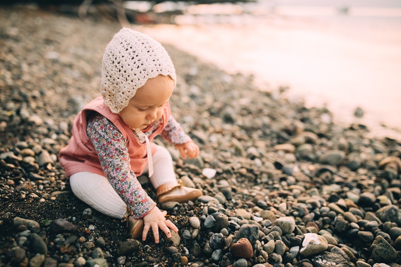 Milestone Photography, a baby girl plays with stones curiously near the lake shore