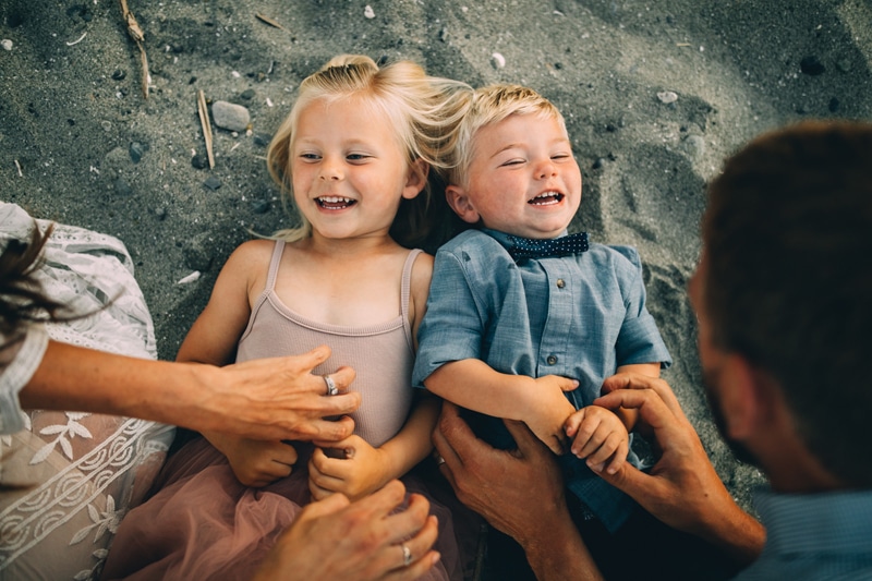 Family Photographer, a young toddler boy and girl lay in the sand as their parents tickle them