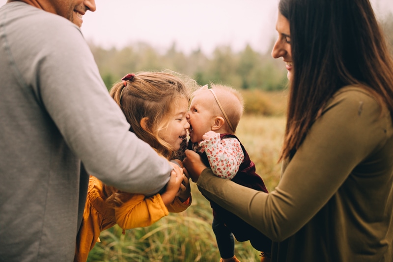 Family photographer, a mom and dad each hold one of their daughters close for kisses as they giggle