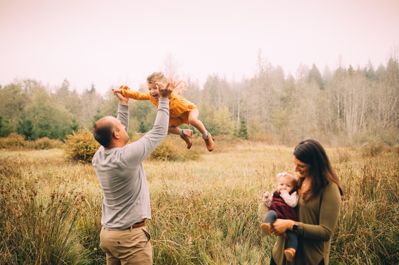 Family Photographer, a father lifts his daughter into the ai, mom holds baby boy next to them in the forest