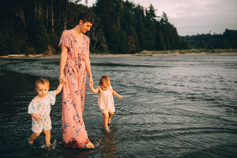 Family Photographer, a woman in a long floral dresses holds her daughters hands as they walk on the lakeshore