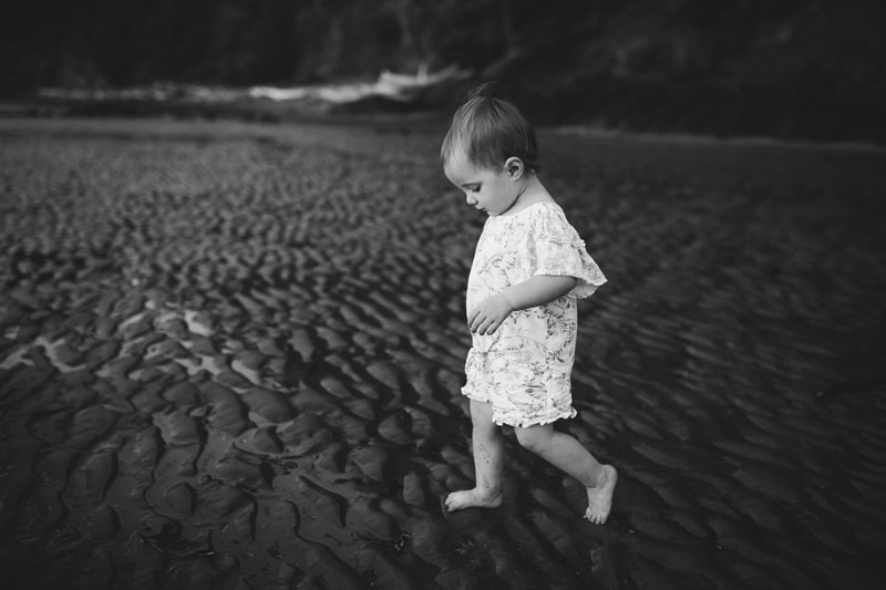 Family photographer, a young toddler girl walks through the sand barefoot