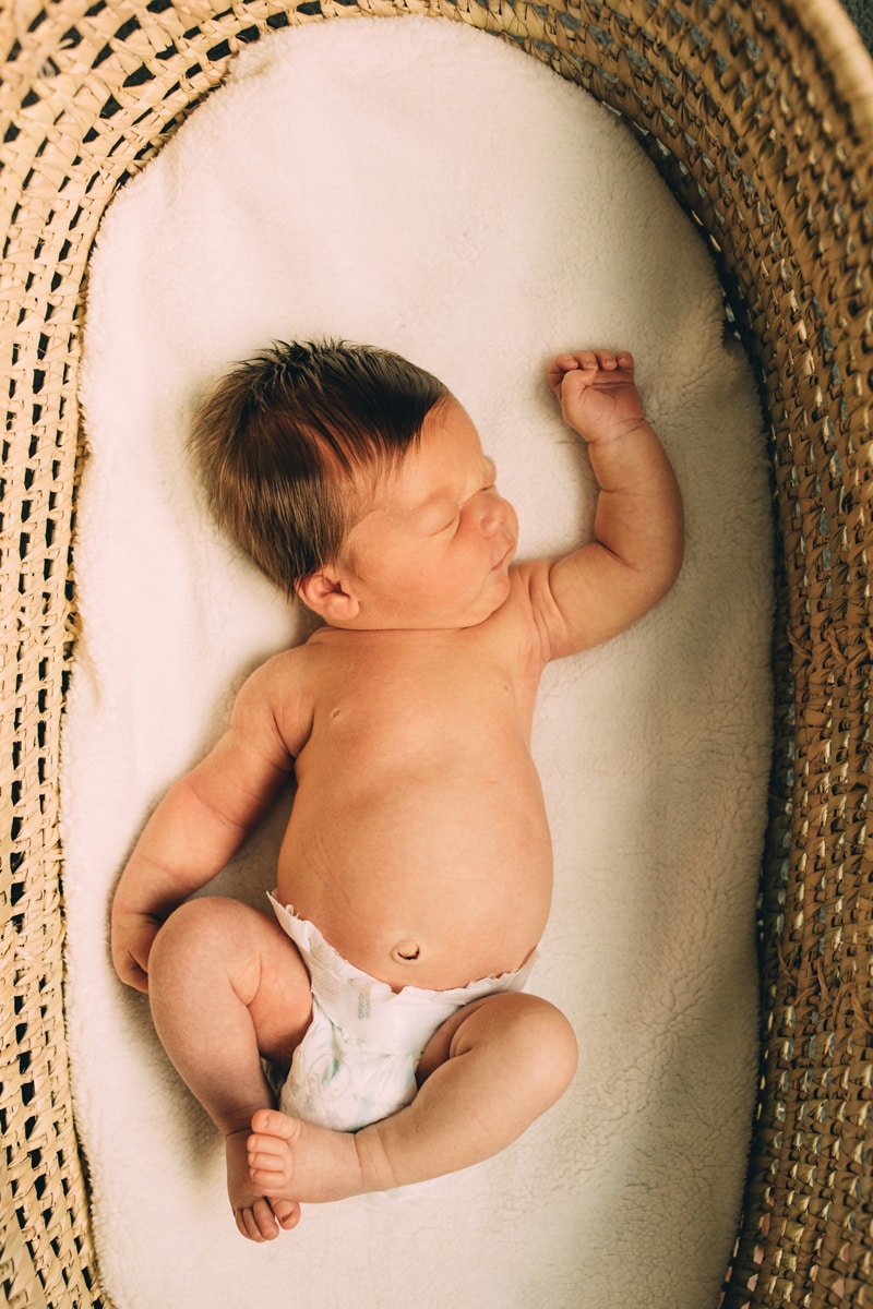 Fresh 48 Newborn Photography, a baby lays in a woven basket, one arm extended above his head, he's sleeping