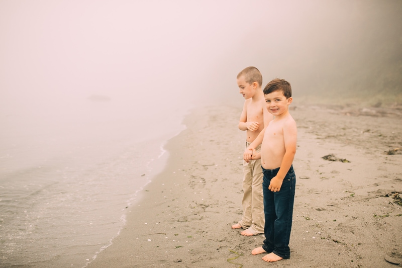 Family Photographer, two young brothers hold hands and smile as they admire the lake on a foggy morning