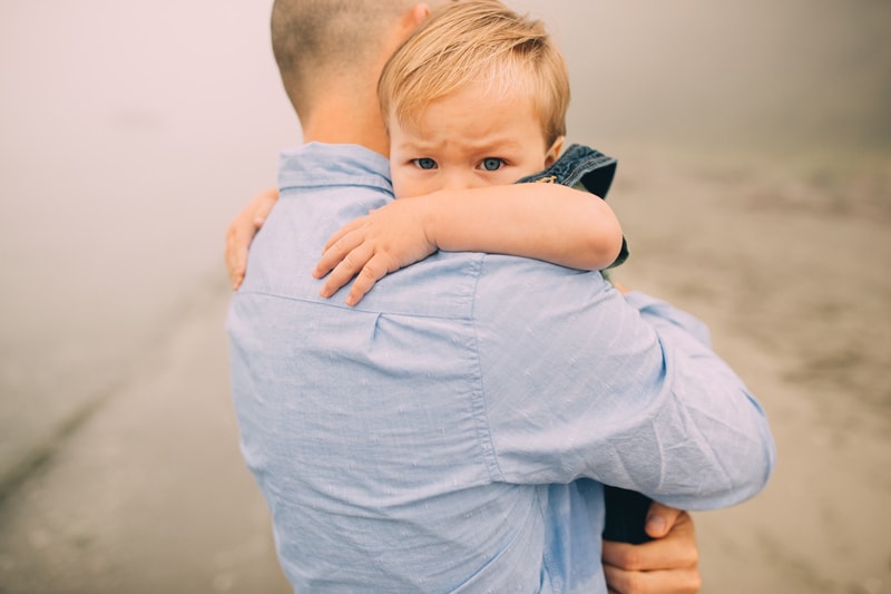 Family photographer, a young boy wraps his arms around his dad's neck as they walk on the beach