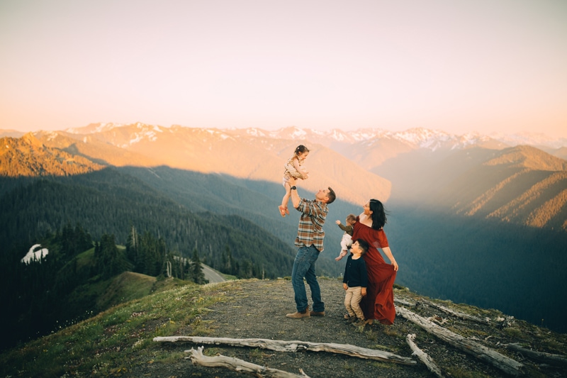 Family Photography, a father lifts baby daughter into the air, mountains behind her. Mom and siblings watch happily