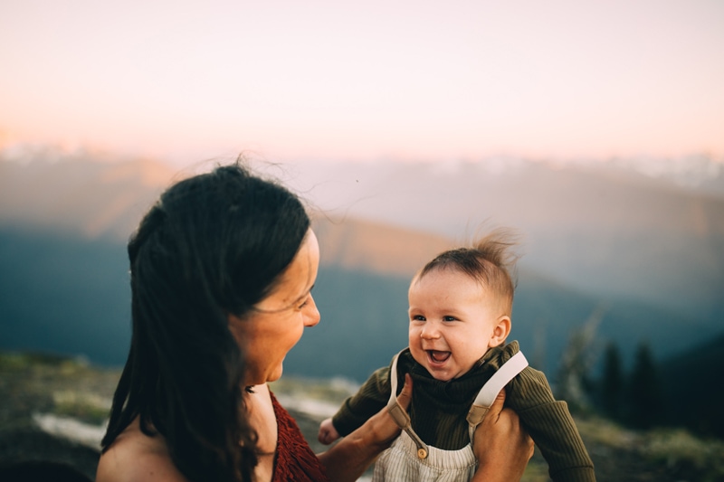 Family photographer, a young mother holds her baby boy up, he is smiling outdoors on a windy evening
