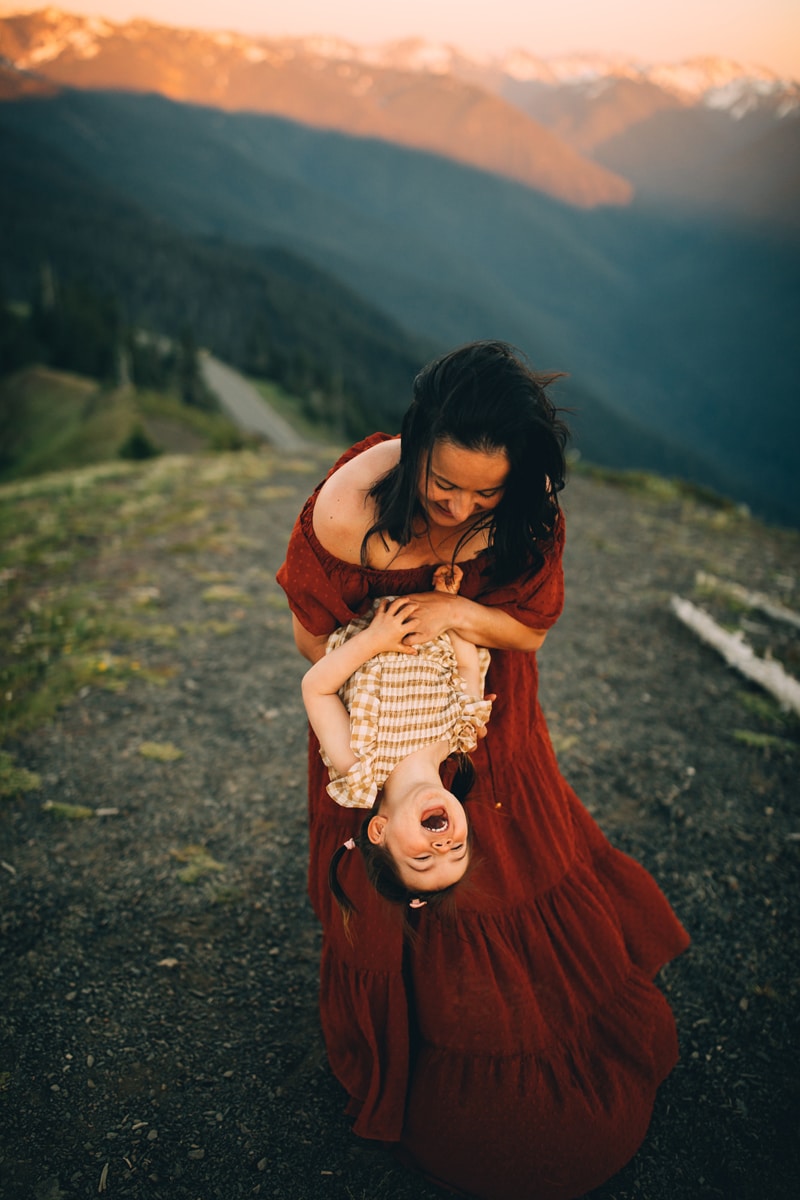 Family Photographer, young mother holds baby girl upside-down playfully outdoors on a hillside