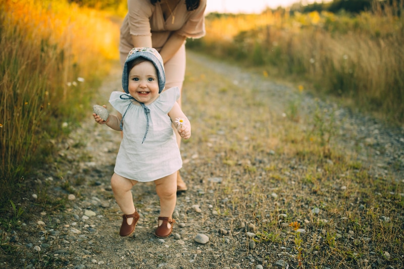 Motherhood Photography, a baby girl wearing a little bonnet walks along a gravel trail through the grass, mom watches on