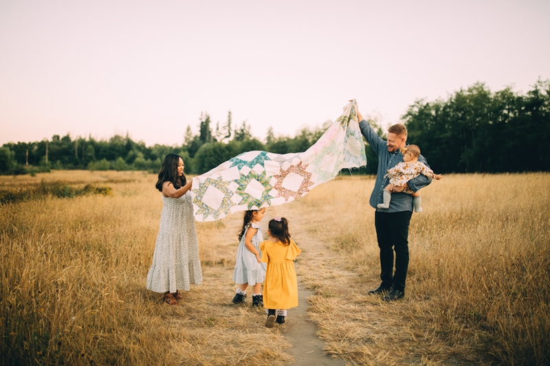 Family Photographer, mom and dad hold up a quilt in afield as their young daugtter run beneath it happy