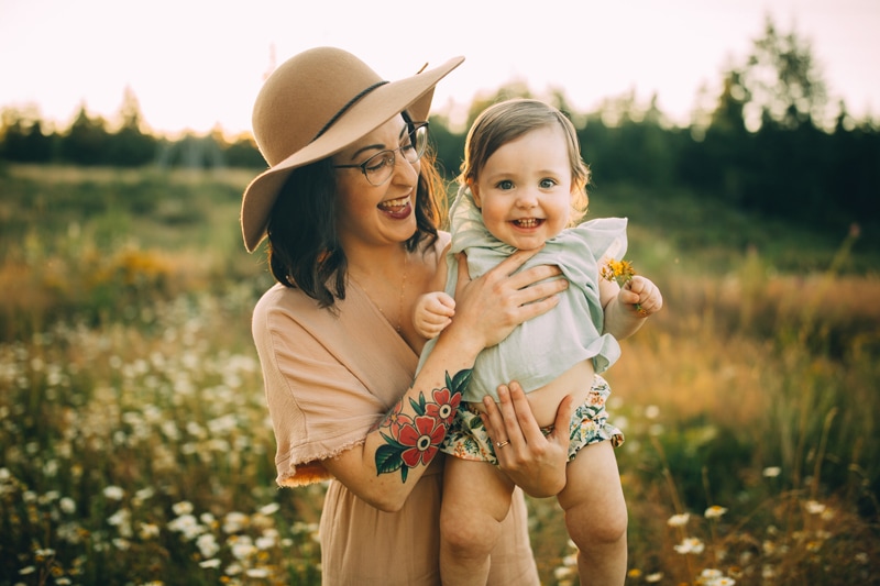 Motherhood Photography, a mom holds her happy toddler daughter as they stand amidst a grass meadow
