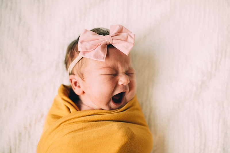 Fresh 48 Newborn Photography, a new baby girls lays tucked in a soft blanket, she has a bow on her full head of hair and yawns