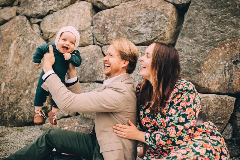 Family photographer, a dad holds up baby smiling, mom leans in also, they sit near large rocks outside