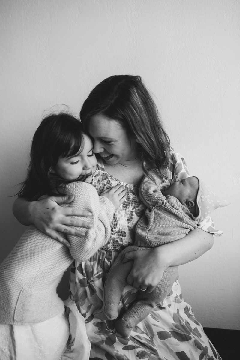 Motherhood photography, mom hugs her toddler daughter and holds newborn baby tight