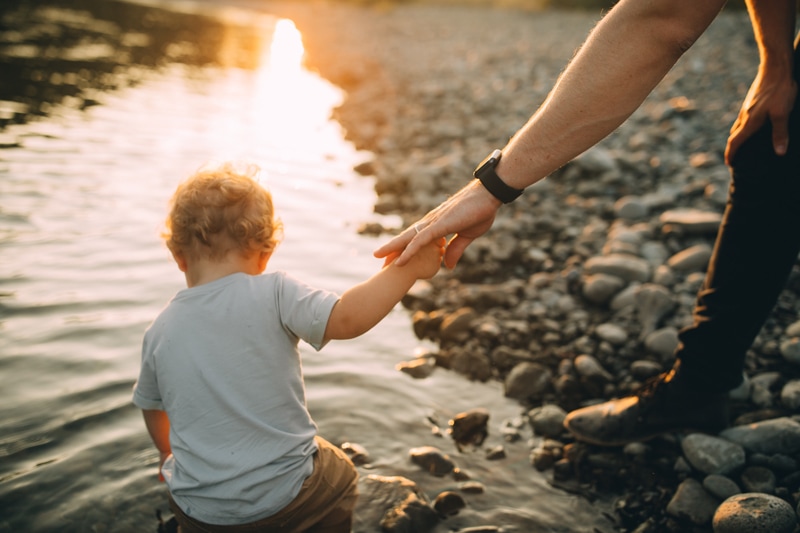 Family photographer, a baby boy sits and plays in the lakeshore, dad holds his hand