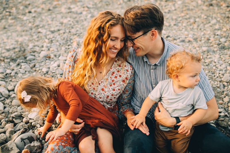 Family photographer, a young man and woman lean into each other happily, they hold their small children in their arms outside