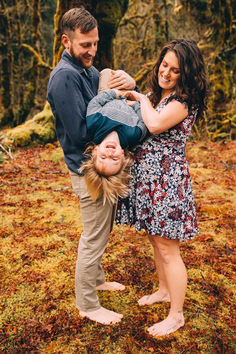 Family photographer, mom and dad hold their young daughter upside down playfully in the forest
