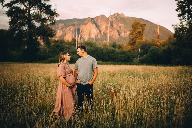 Family Photographer, a man and woman gaze into each other's eyes content in a meadow, she holds her belly as they are expecting baby