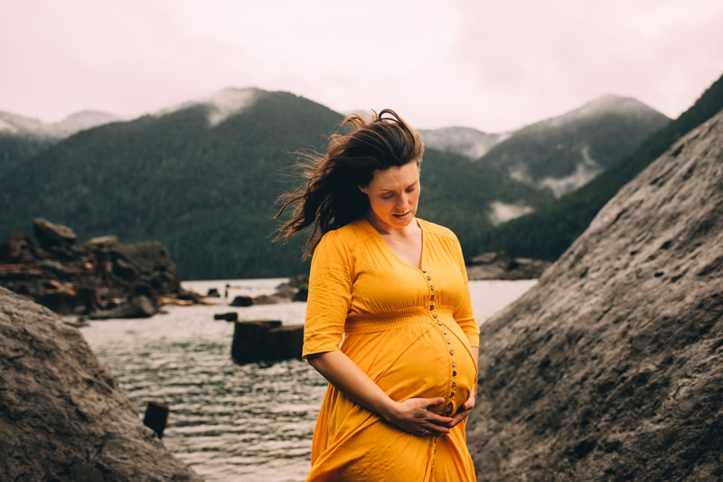 Family photographer, a woman holds her belly near a lakeside, she is expecting