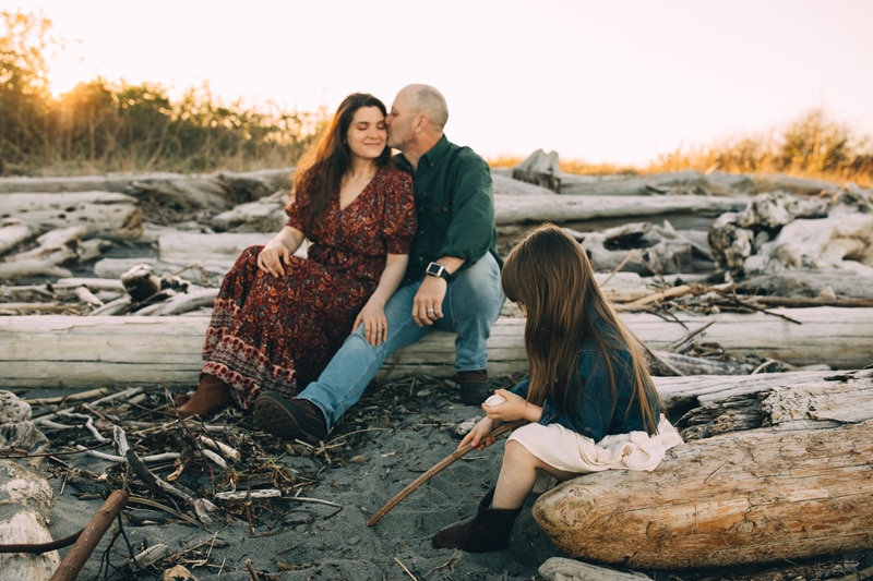 Family photographer, a man holds his wife and kisses her on the head, their daughter sits on a log near to them