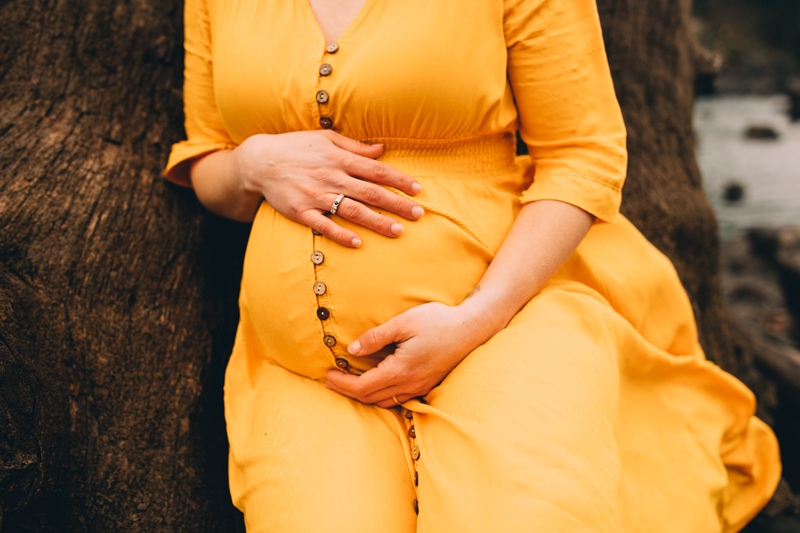 Family Photographer, a woman holds her belly as she leans against a tree, she is pregnant