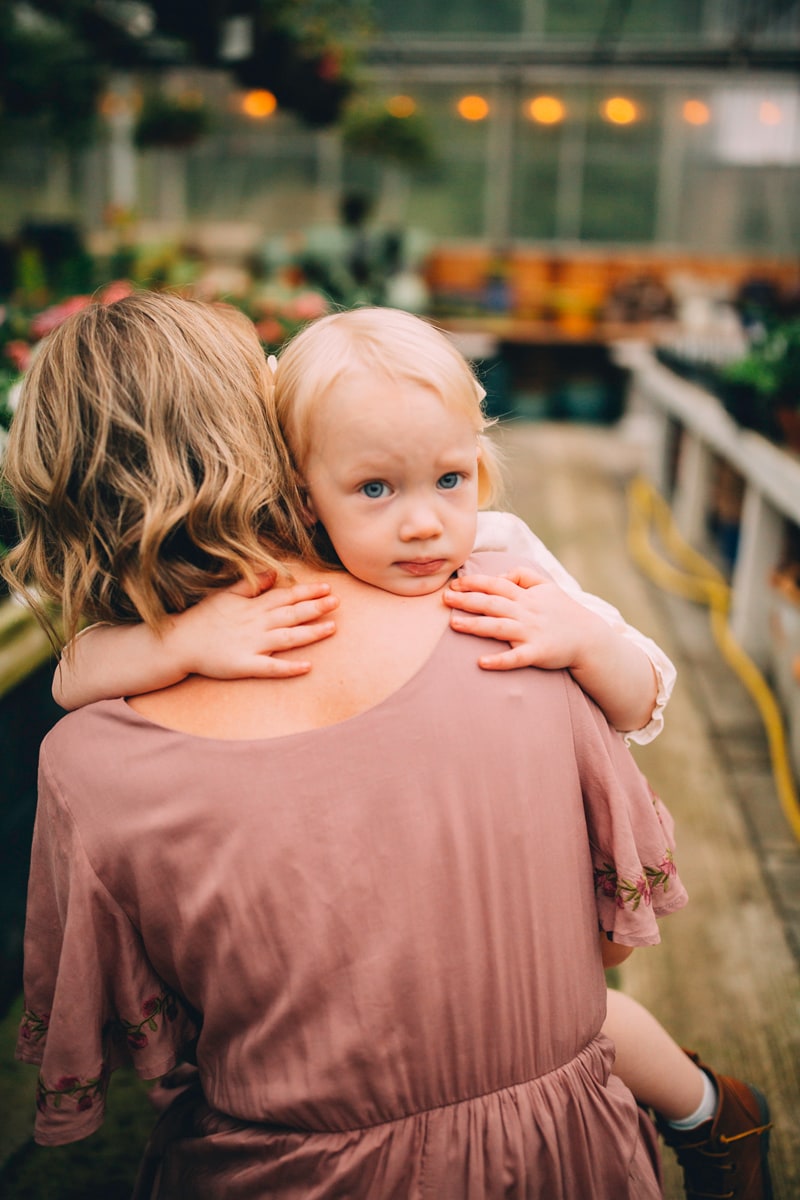 Motherhood photography, a little girl squeezes mom's she is carried through a plant nursery