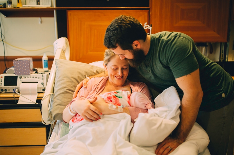 Fresh 48 Newborn Photography, a mother lays in a hospital recovery bed while holding their baby daughter, dad leans in to dote as well