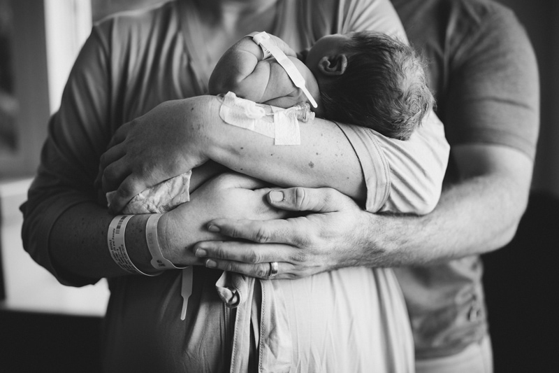 Fresh 48 Newborn Photography, a new mother holds her baby in her arms, dad holds mom and admires baby from behind