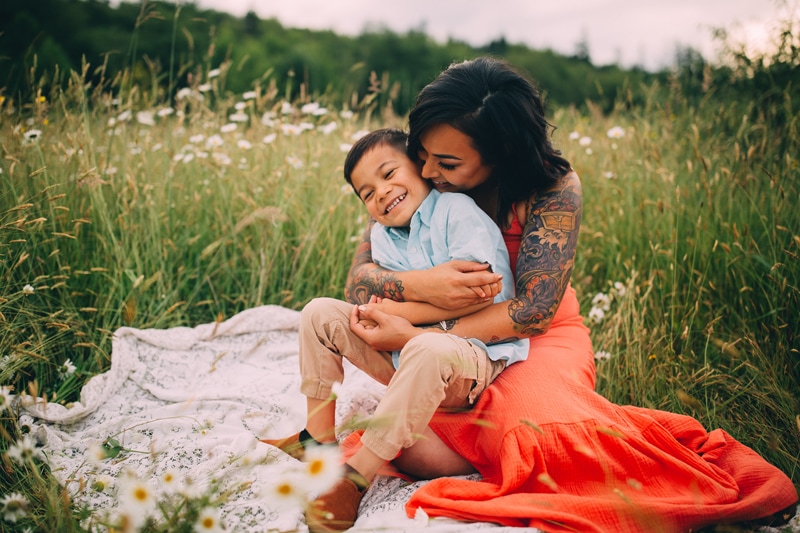 Motherhood Photography, a young mother with tattoos on her arms squeezes her little boy tight wit h love on a blanket in a grassy meadow