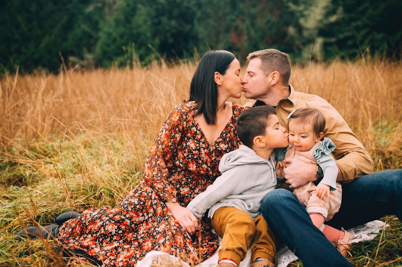 Family Photographer, Mom and dad kiss on a blanket in the a meadow. They hold their two little children.