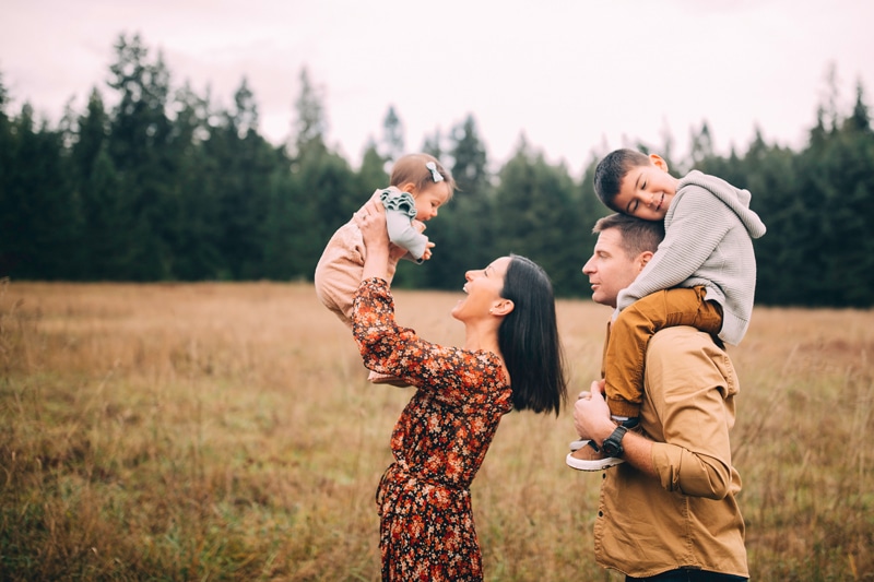 Family Photographer - Mom holds up baby with a big smile, older brother sits on dad's shoulders besides them , they are in open grass field